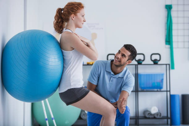 uśmiechnięty profesjonalny trener personalny pomagający sportsmence ćwiczącej z piłką - adult blue caucasian doctor zdjęcia i obrazy z banku zdjęć