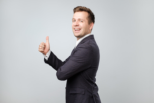 Portrait of excited man in suit giving thumb up against gray background. He is approving the choice. It is cool