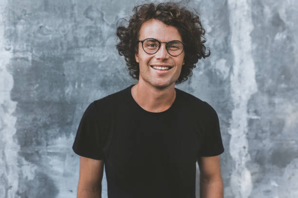 Outdoor portrait of handsome freckled smiling male with curly hair, wears sepctacles posing for social advertisement, isolated on gray concrete wall with copy space for your promotional information stock photo