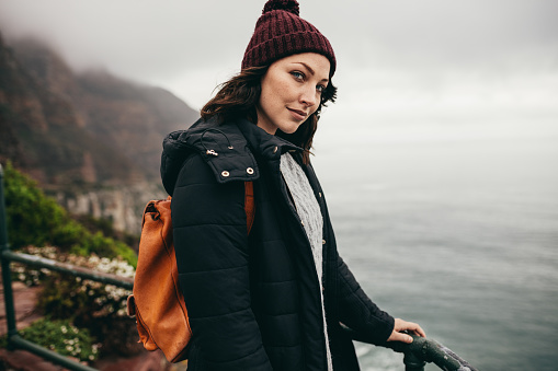Portrait of beautiful young woman standing by a railing at hill top and looking at camera. Female on vacation wearing warm clothing and carrying a bag.