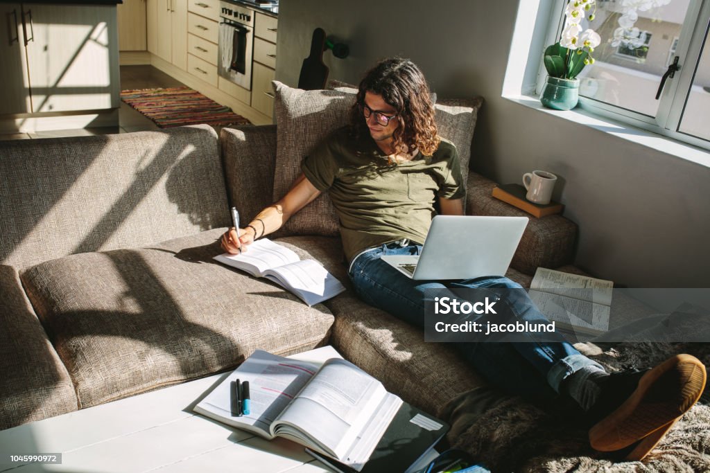 Student studying at home sitting beside a window Student studying for exams sitting at home comfortably on a couch. Young man using laptop to study along with books. E-Learning Stock Photo