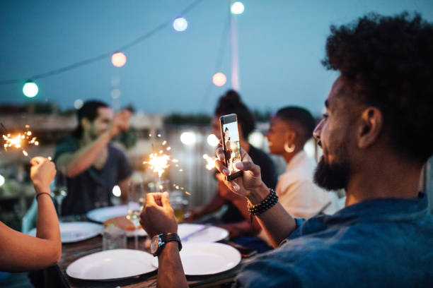 Young man taking picture of sparkler at rooftop party Young man taking picture of sparkler with friends sitting at table during party in evening. People sitting at rooftop enjoying the party. berlin photos stock pictures, royalty-free photos & images