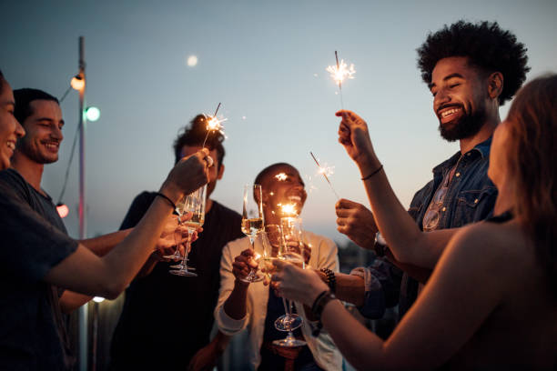 Friends celebrating at party on rooftop Multiracial young men and women on roof raising wineglasses and sparklers. Friends celebrating at reunion party on rooftop. reunion social gathering stock pictures, royalty-free photos & images
