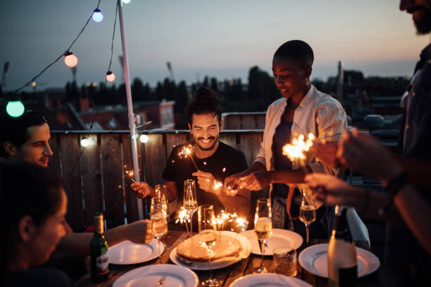 Multi-ethnic friends enjoying rooftop party Multi-ethnic friends sitting around a table with cake and sparklers in hand at rooftop party.  Men and women igniting sparklers placed on a cake during sunset. people banque stock pictures, royalty-free photos & images