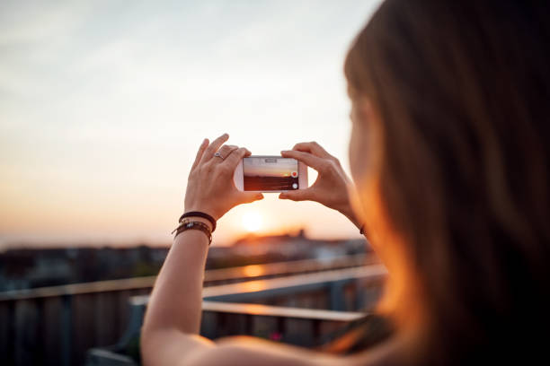 Woman taking pictures of sunset from rooftop Rear view of woman shooting a photograph of sunset with her mobile phone while standing on roof of a building. building terrace photos stock pictures, royalty-free photos & images