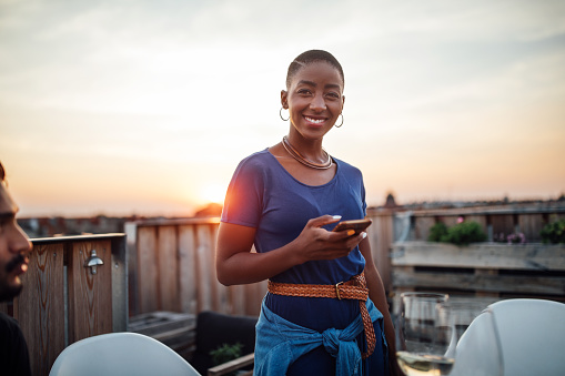 Beautiful young african woman with bald head standing on roof holding her mobile phone. Female at rooftop party with phone at sunset.