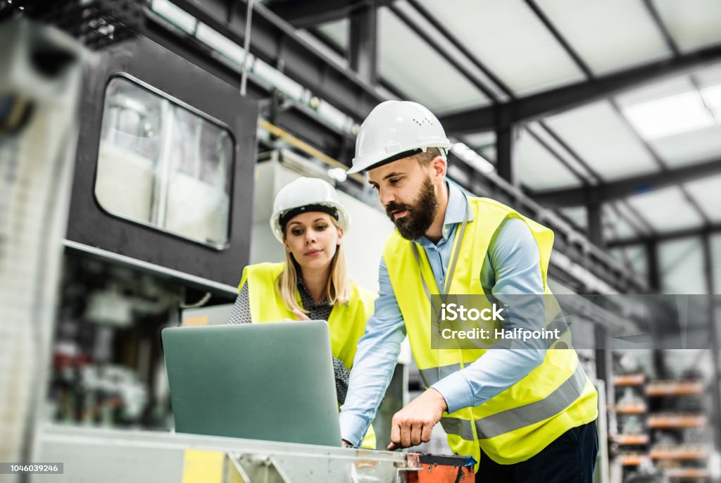 Un retrato del ingeniero industrial del hombre y la mujer con el portátil en una fábrica, trabajando. - Foto de stock de Manufacturar libre de derechos