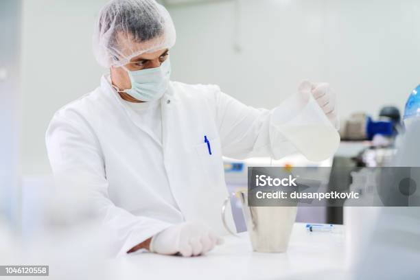 Picture Of Man In Sterile Clothes In Bright Laboratory Pouring Transparent Liquid In Dish Experiment In Process Stock Photo - Download Image Now
