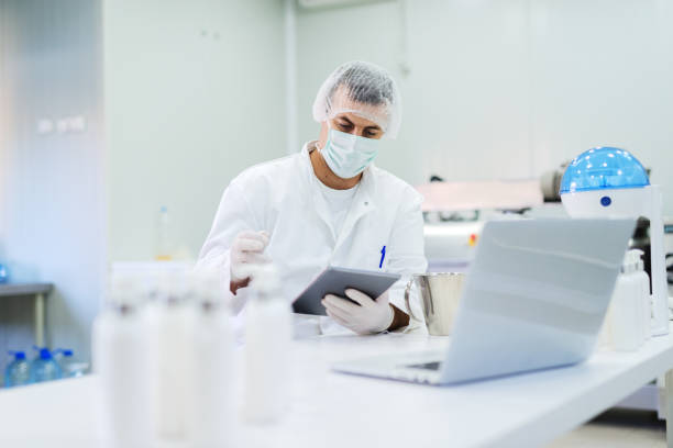 Picture of man in sterile clothes sitting in bright laboratory and checking quality of products. Holding tablet in his hands and reading notes. Picture of man in sterile clothes sitting in bright laboratory and checking quality of products. Holding tablet in his hands and reading notes. pharmaceutical factory stock pictures, royalty-free photos & images