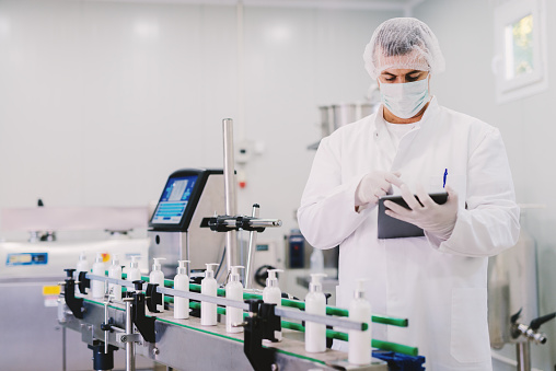 Picture of young man in sterile clothes using tablet . Controlling production line on factory. Bottles with cosmetic products on production line.