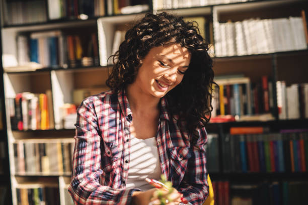 istock Beautiful charming young student girl sitting in library and learning. Smiling and taking notes. 1046124772