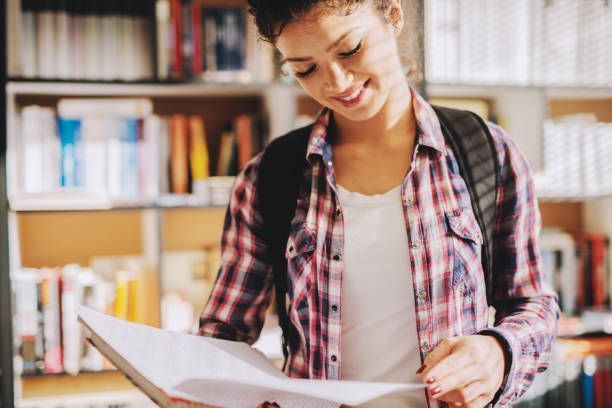 istock Beautiful smiling young school girl with backpack on her back standing in bright library and looking at book. Blurred picture of bookshelves in background. 1046128066