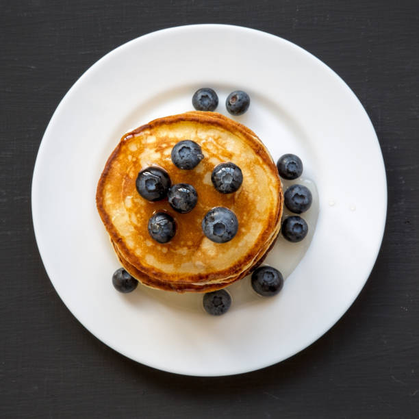 Pancakes with blueberries and honey on white plate on dark wooden table, overhead view. Flat lay, top view, from above.
