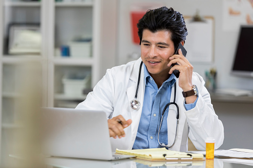 Hispanic male doctor talks with patient on the phone. He is looking at a laptop while talking with the patient.