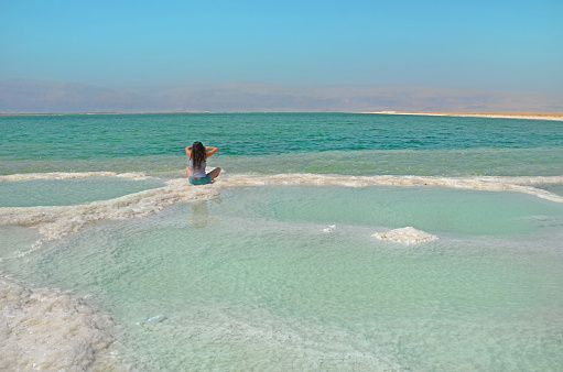 long-haired brunette woman sitting on a path of salt in the sea. The water surface of the Dead Sea in Israel with a view of the mountains of Jordan. Merging with nature.