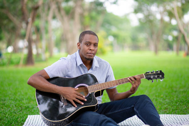 homme noir jouant de la guitare et assis sur le sol dans le parc - musical instrument nature outdoors musician photos et images de collection