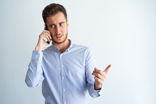 Doubtful businessman pointing finger away. Young man in formal shirt talking on cell and showing copy space near him. Communication concept