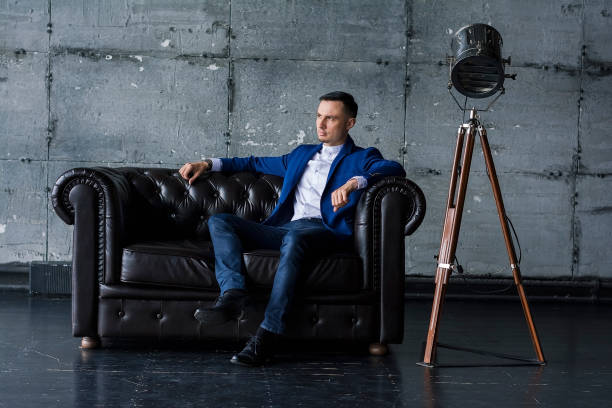 istock Beautiful elegant fashionable young man in a blue jacket sits on a leather sofa 1046451126