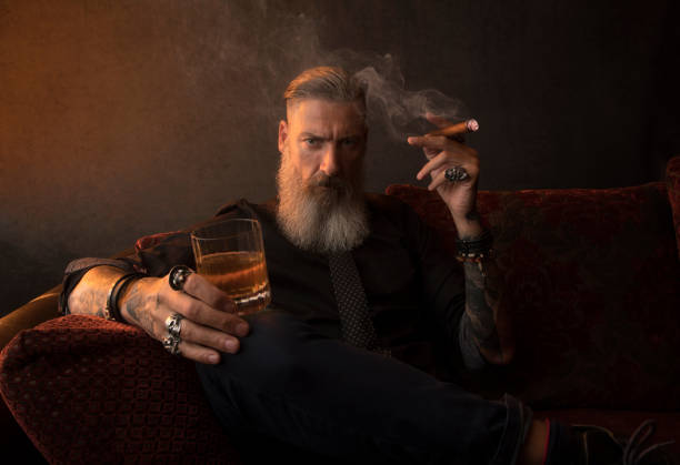 portrait d’un homme d’affaires attrayant avec un cigare et un verre de whisky dans une pièce sombre - orange beauty serious retro revival photos et images de collection