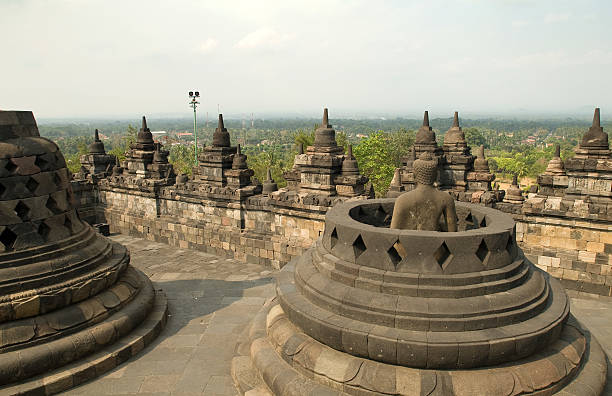 Photograph of a Buddha statue of buddha in one of the stupas on top of the Borobudur ruins in Jogjakarta, Java Island, Indonesia, overlooking the land. The Borobudur stupa with Buddhist and Saivist elements, derived from Brahminism dates to the ninth century A.D.