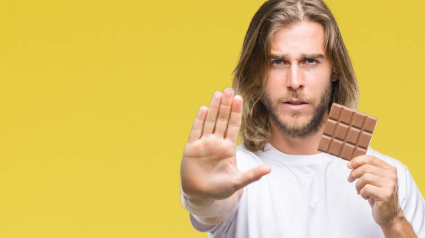 istock Young handsome man with long hair eating chocolate bar over isolated background with open hand doing stop sign with serious and confident expression, defense gesture 1046560310