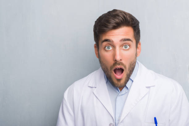 istock Handsome young professional man over grey grunge wall wearing white coat afraid and shocked with surprise expression, fear and excited face. 1046563508