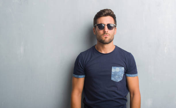 istock Handsome young man over grey grunge wall wearing sunglasses with serious expression on face. Simple and natural looking at the camera. 1046565622