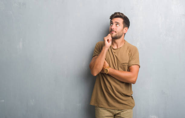istock Handsome young man over grey grunge wall with hand on chin thinking about question, pensive expression. Smiling with thoughtful face. Doubt concept. 1046565726