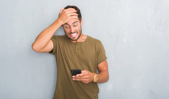 Handsome young man over grey grunge wall texting a message using smartphone stressed with hand on head, shocked with shame and surprise face, angry and frustrated. Fear and upset for mistake.