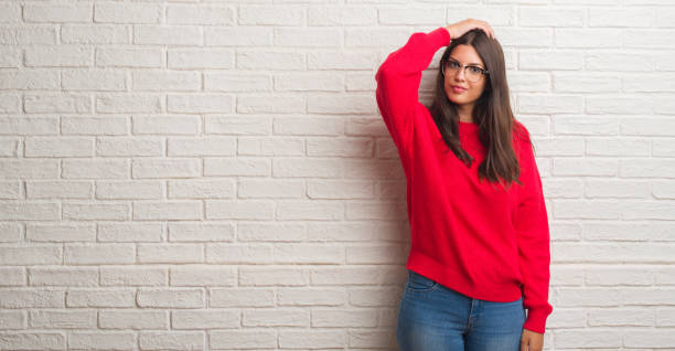 Young brunette woman standing over white brick wall confuse and wonder about question. Uncertain with doubt, thinking with hand on head. Pensive concept.