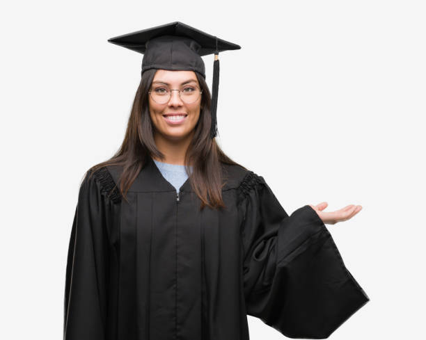 istock Young hispanic woman wearing graduated cap and uniform smiling cheerful presenting and pointing with palm of hand looking at the camera. 1046570568