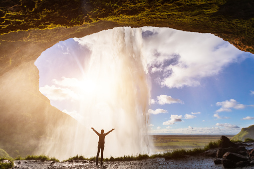 Happy female traveler feeling the power of Seljalandsfoss waterfall in the South of Iceland, person standing behind the stream, sunset light, intense icelandic experience