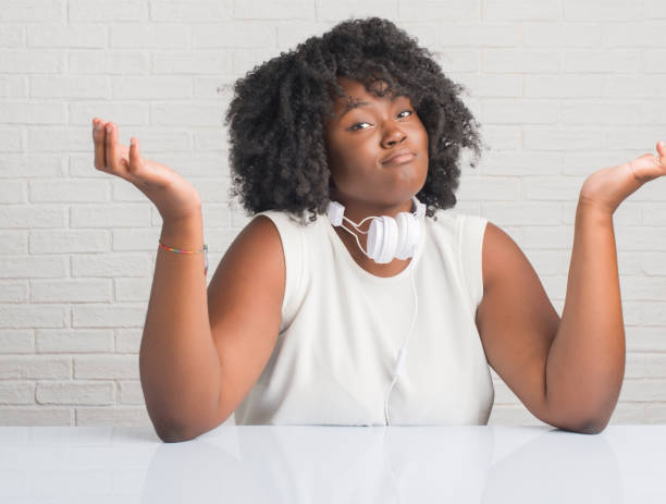 young african american woman sitting on the table wearing headphones clueless and confused expression with arms and hands raised. doubt concept. - head and shoulders audio imagens e fotografias de stock