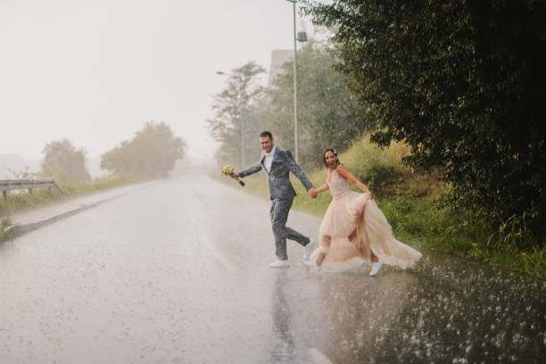 tola jovem casal recém casado cruzamento estrada em dia chuvoso. correndo em roupas molhadas cerimoniais. - wet dress rain clothing - fotografias e filmes do acervo