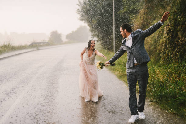 totalmente molhada só casal andando na chuva pela estrada de carro. noiva é treinar para pegar o marido. - wet dress rain clothing - fotografias e filmes do acervo