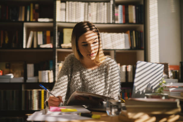 Cute young school girl sitting in library and taking notes while looking at laptop. Sitting alone and studying in bright library.