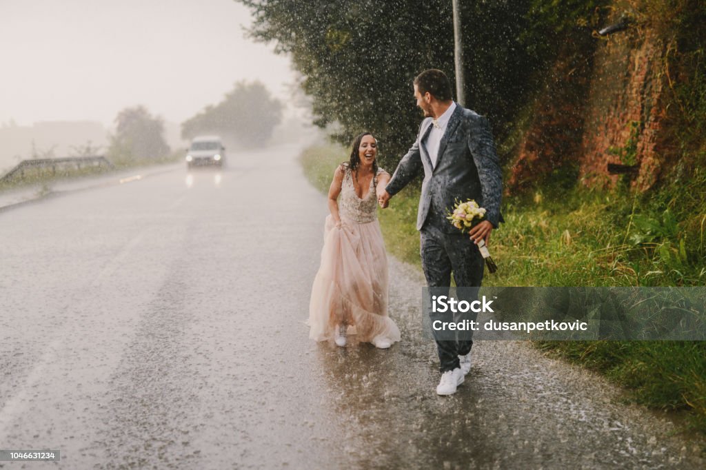 Couple juste marié tenant par la main et de marcher sur la pluie. Marcher dans des vêtements de cérémonie mouillés sur route en voiture. Souriant et s’amusant. - Photo de Mariage libre de droits