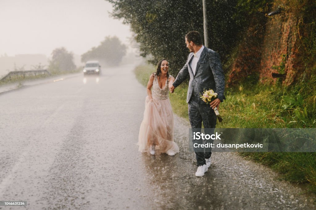 Coppia appena sposata che si tiene per mano e cammina sotto la pioggia. Camminare in abiti cerimoniali bagnati sulla strada di guida. Sorridere e divertirsi. - Foto stock royalty-free di Matrimonio