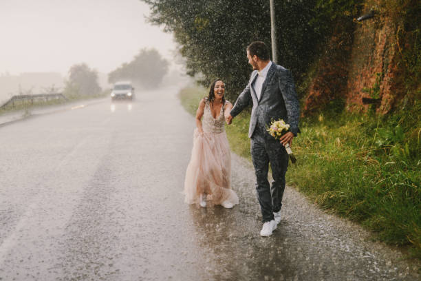 ちょうど結婚されていたカップルの手を繋いでいると雨の上を歩きます。ドライブの道に濡れた儀式服を着て歩いています。笑顔と楽しい時を過します。 - wet dress rain clothing ストックフォトと画像