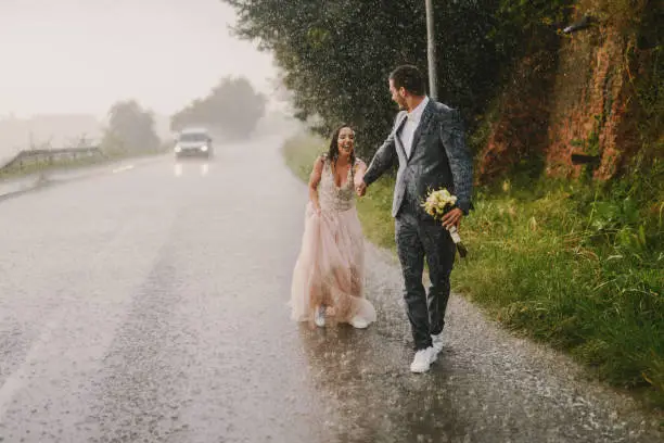 Photo of Just married couple holding hands and walking on rain. Walking in wet ceremonial clothes on drive road. Smiling and having fun.