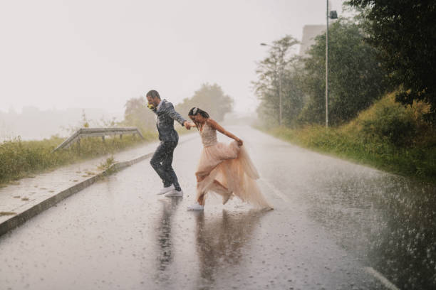 tola jovem casal recém casado cruzamento estrada em dia chuvoso. correndo em roupas molhadas cerimoniais. - wet dress rain clothing - fotografias e filmes do acervo