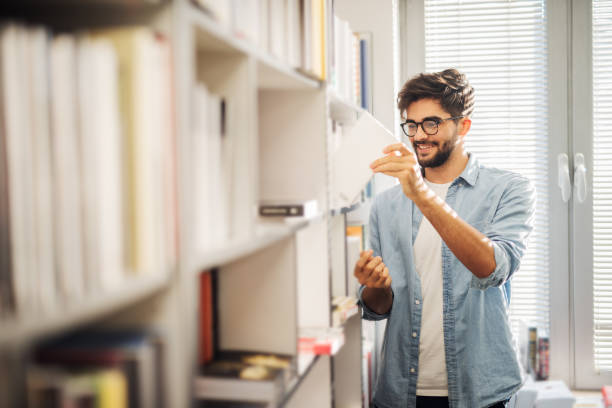 mignon jeune urbain étudiant debout dans la bibliothèque vif et prenant livre de bibliothèque. seuls et souriant. - library young adult bookstore people photos et images de collection