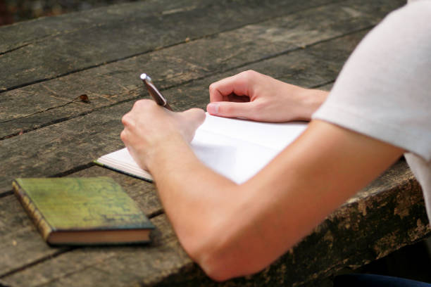istock Young handsome guy sitting at wooden table, writing a book, doing homework, taking notes, learning, contemplating and writing down his thoughts and lessons while enjoying the outdoors in forest. 1046738238