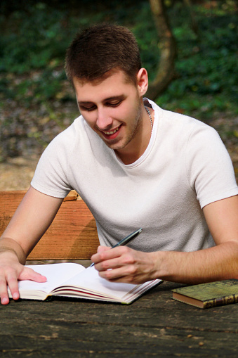 Guy and student takes notes in notebook, learning and writes thoughts, writes the book, he is preparing his final exams doing homework and thinking. Man is writing something in his notebook in forest.