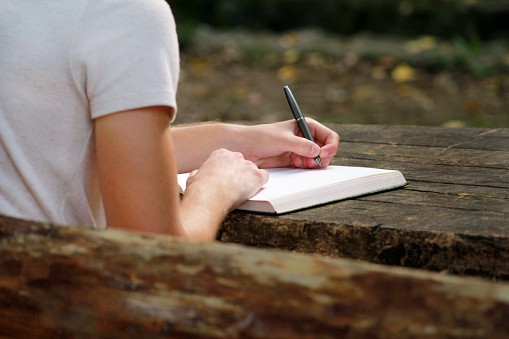 Young handsome guy sitting at wooden table, writing a book, doing homework, taking notes, learning, contemplating and writing down his thoughts and lessons while enjoying the outdoors in forest.