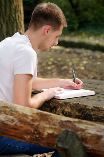 istock Young handsome guy sitting at wooden table, writing a book, doing homework, taking notes, learning, contemplating and writing down his thoughts and lessons while enjoying the outdoors in forest. 1046738364