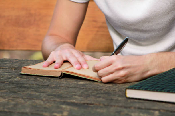 istock Young handsome guy sitting at wooden table, writing a book, doing homework, taking notes, learning, contemplating and writing down his thoughts and lessons while enjoying the outdoors in forest. 1046738388
