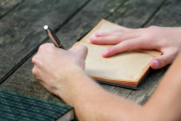 istock Young handsome guy sitting at wooden table, writing a book, doing homework, taking notes, learning, contemplating and writing down his thoughts and lessons while enjoying the outdoors in forest. 1046738394