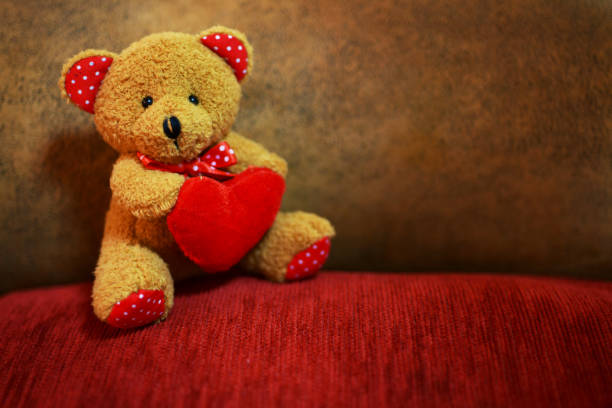 horizontal image of a little soft tean coloured teddy bear surrounded by red and pink hearts on white background.