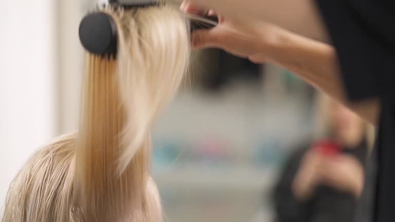 A close up of blonde's hair dried in strands. The hand takes a strand with a round brush, stretches it bottom up and dries along with a hairdryer.